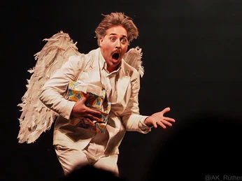 Cirque Bouffon - Célébration Ein Künstler in weißem Anzug und Engelsflügeln, mit ausdrucksstarker Mimik und offenem Mund.An artist in a white suit and angel wings, with expressive facial expressions and an open mouth.