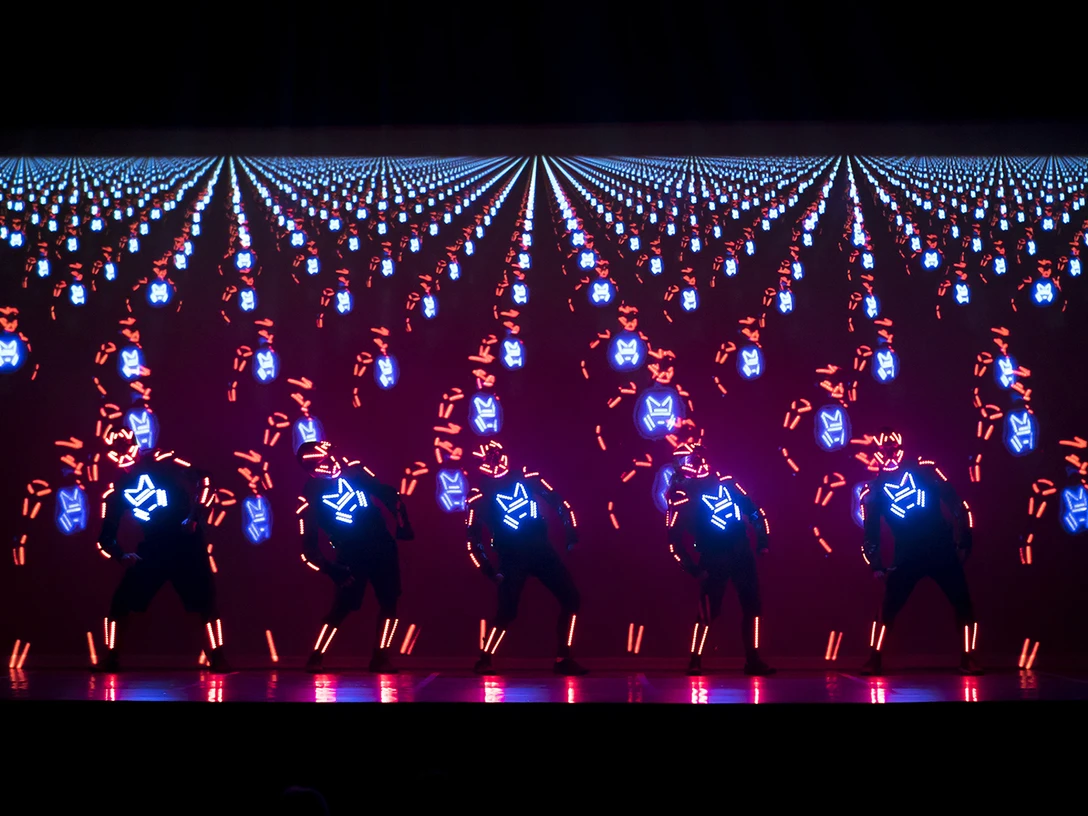 Brodas Bros_AROUND THE WORLD.jpg Das Bild zeigt eine Tanzformation in schwarzer Kleidung mit neonfarbenen Mustern auf der Vorderseite. Sie tanzen im Lichtschein