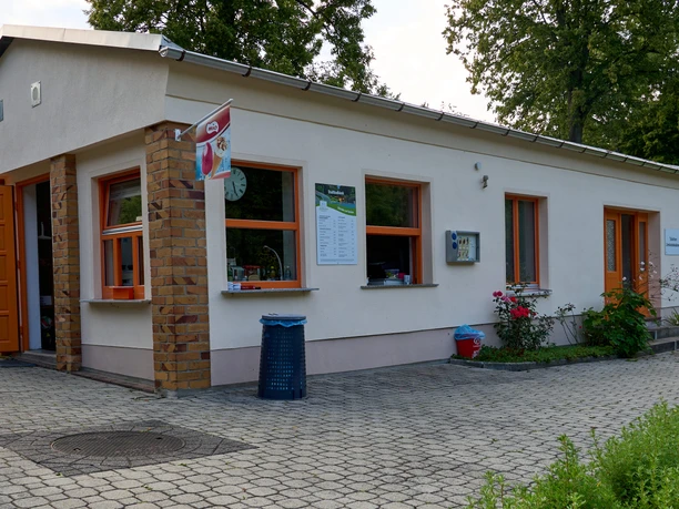 Malá bílá budova s oranžovými dveřmi a okenními rámy, obklopená zelení.