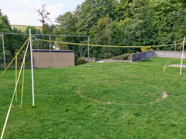 Leerer Volleyballplatz auf grüner Wiese mit Netz und Metallzaun im Hintergrund.