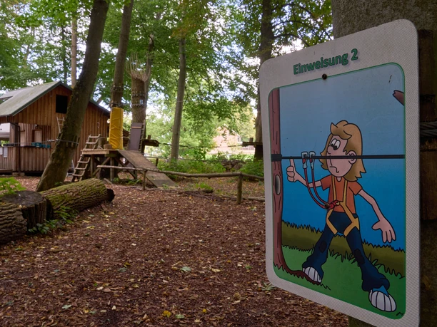 Sign with balancing exercise in the climbing forest, wooden hut in the background.