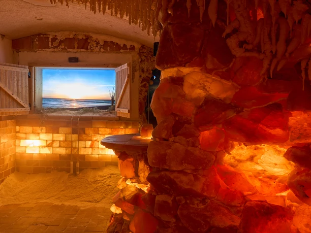 Salzgrotte Lindenhof Königstein Innenraum einer Salzgrotte mit beleuchteten Salzsteinwänden und TV-Fensterblick auf den Sonnenuntergang am Meer.