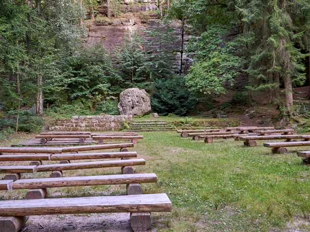 Openluchtpodium in het bos met houten banken en een rotswand op de achtergrond.