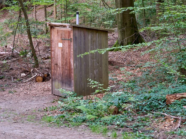 Houten toilet in het bos naast een pad, omringd door bomen en struiken.