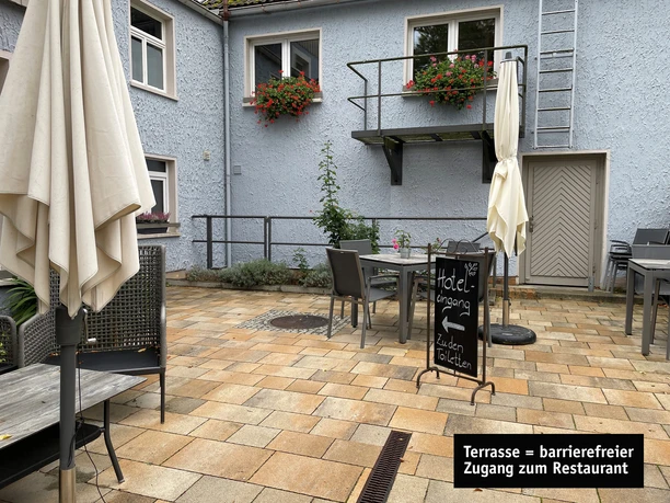 Eingang und Terrasse.jpg Terrasse mit rotgelbem Pflaster, Tischen und Stühlen, Sonnenschirmen und Fenstern mit Blumenkästen