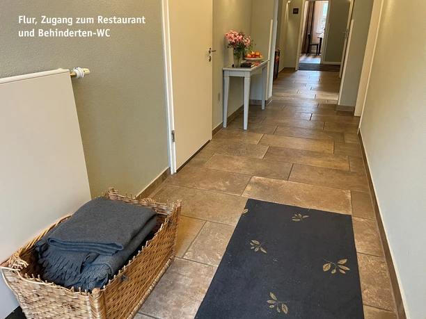 Koridor.jpg Široká chodba s červenohnědou dlažbou, černým běhounem, košem plným dek, malým stolkem s květinovou výzdobou a několika bílými dveřmi.