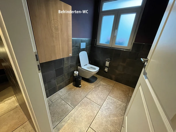 Behinderten-WC.jpg Toilettenraum mit Wickeltisch an der Wand, WC Becken und großzügigem Fenster