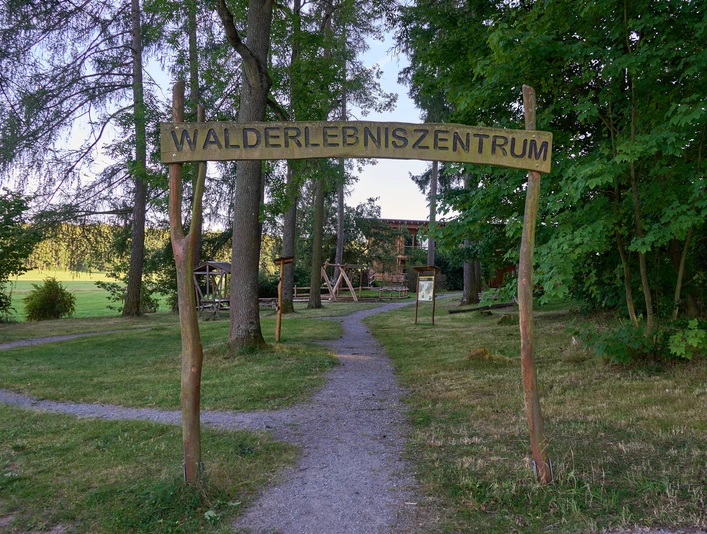 Walderlebniszentrum Holzschild "Walderlebniszentrum" über einem Weg, der in einen Park führt.Wooden sign "Forest Experience Center" above a path leading into a park.Dřevěná cedule "Forest Experience Centre" nad cestou vedoucí do parku.Drewniany znak "Forest Experience Centre" nad ścieżką prowadzącą do parku.Houten bord "Forest Experience Centre" boven een pad dat naar een park leidt.Cartello in legno "Forest Experience Centre" sopra un sentiero che conduce a un parco.