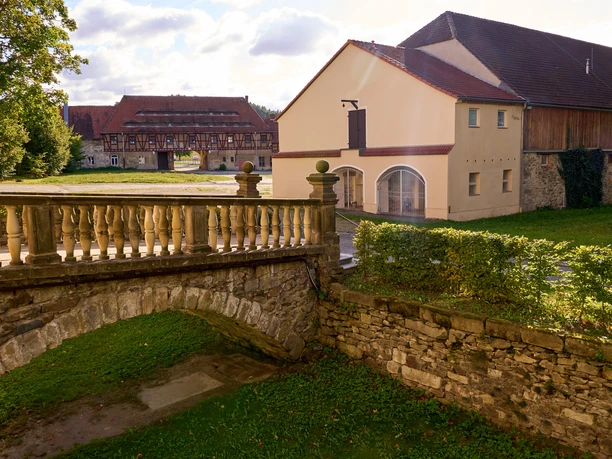 Kulturscheune und Schloss Langburkersdorf Steinbrücke mit Geländer vor historischen Gebäuden und grüner Wiese.