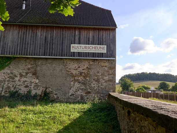 Stenen muur op de voorgrond, daarachter schuur met bord "KULTURSCHEUNE", landschap op de achtergrond.