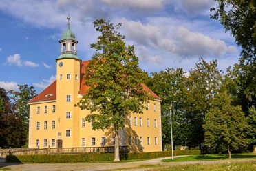 Kulturscheune Schloss Langburkersdorf Gelbes Schloss mit grünem Dach und Turm, umgeben von Bäumen unter blauem Himmel.