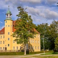 Kulturscheune Schloss Langburkersdorf Gelbes Schloss mit grünem Dach und Turm, umgeben von Bäumen unter blauem Himmel.