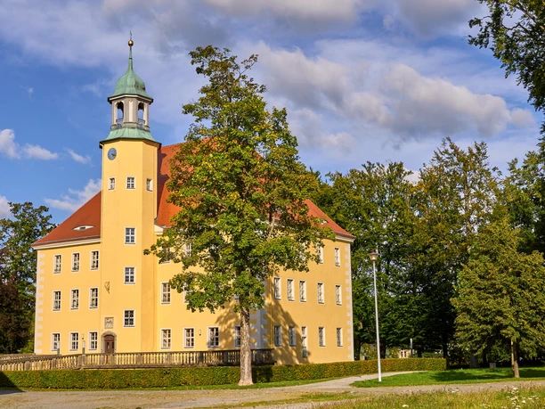 Kulturscheune Schloss Langburkersdorf Gelbes Schloss mit grünem Dach und Turm, umgeben von Bäumen unter blauem Himmel.