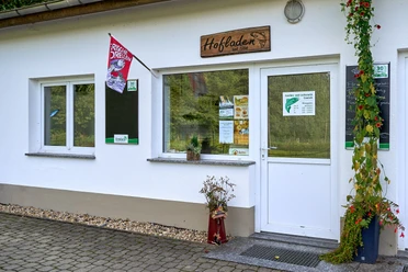 Wit gebouw met uithangbord "Hofladen", toegangsdeur en twee ramen, versierd met planten.