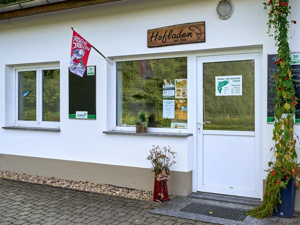 White building with "Hofladen" sign, entrance door and two windows, decorated with plants.