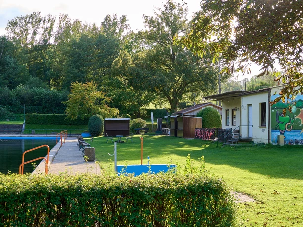 Waldbad Freibad mit grünem Rasen und Graffiti an einer Wand, umgeben von Bäumen.