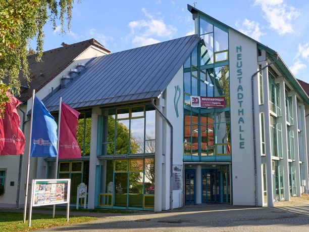 Moderní skleněná fasáda Neustadthalle s modrou střechou a vlajkami.