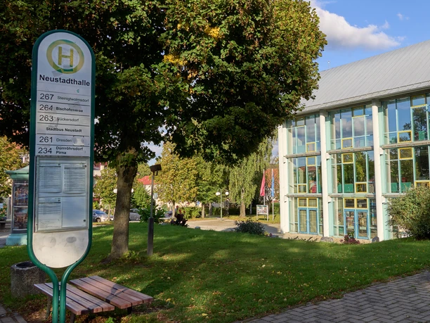Bus stop with timetable in front of a building with a large glass façade and trees.