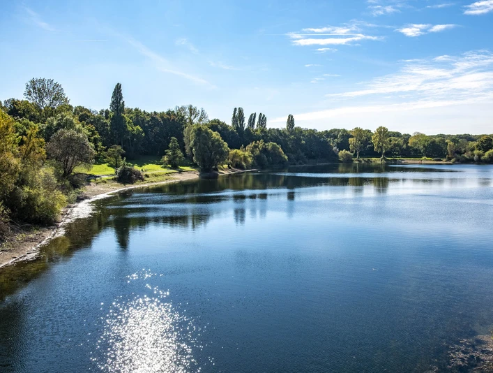 Lake Fühlingen Ein klarer See mit reflektierendem Sonnenlicht und umliegendem Grün unter blauem Himmel in Köln.A clear lake with reflecting sunlight and surrounding greenery under a blue sky in Cologne.