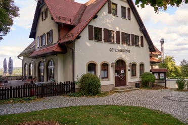 White building with brown roof and "Götzinger Höhe" lettering, surrounded by trees.