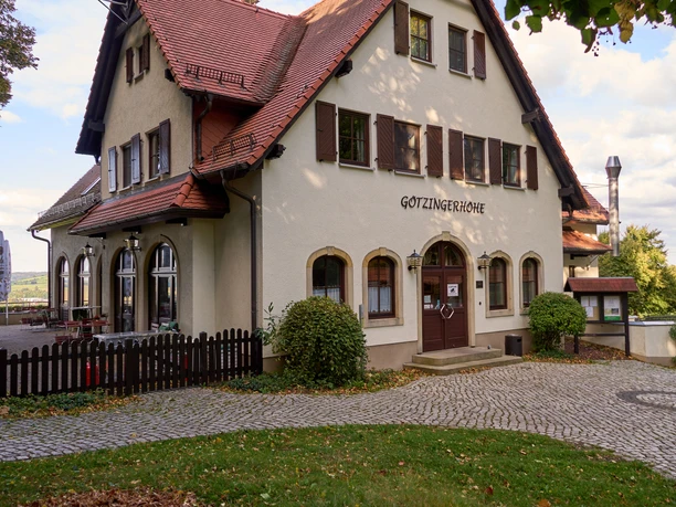 Bílá budova s hnědou střechou a nápisem "Götzinger Höhe", obklopená stromy.