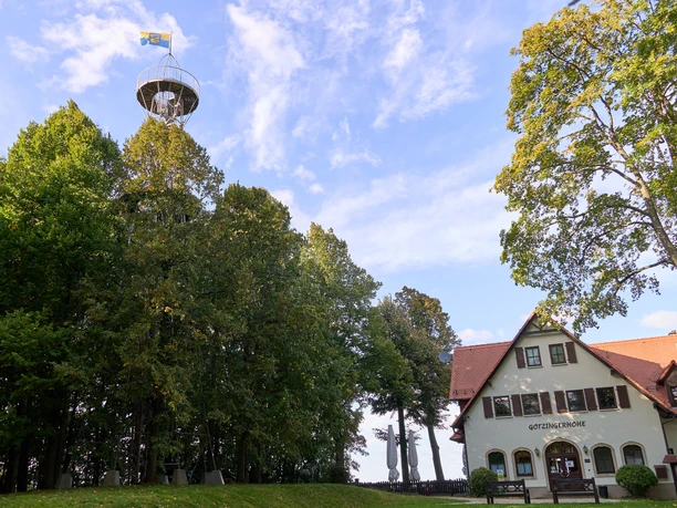 Uitkijktoren achter bomen naast een huis met bord "Götzinger Höhe".