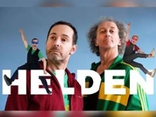 Zwei Männer in farbenfroher Kleidung posieren vor blauem Hintergrund mit "HELDEN"-Schriftzug.