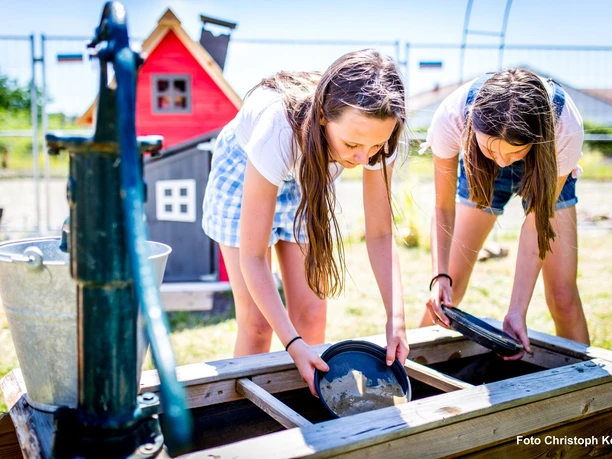 Edelsteinbrunnen im Happylino-Außenbereich Zwei Kinder sieben mit Schalen nach Edelsteinen an einem Wasserbrunnen bei sonnigem Wetter im Happylino Außenbereich.