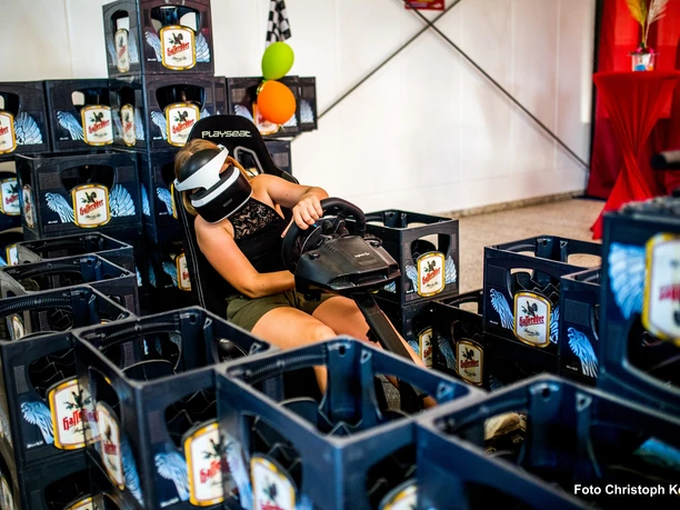 VR-Challenge im Happylino Person in VR-Brille fährt im Rennsimulator, umgeben von leeren Getränkekästen in Rennstrecke-Form.