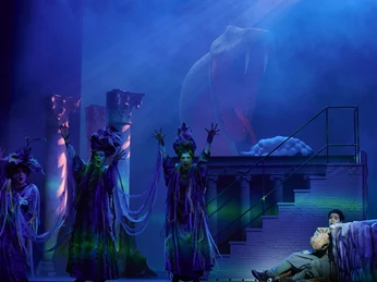 Zauberflöte_2_Foto_Matthias Baus.jpg Vor einer dunklen Bühnenkulisse stehen kostümierte Operndarsteller.