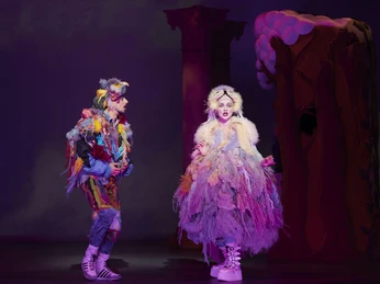 Zauberflöte_1_Foto_Matthias Baus.jpg Vor einer bunten Bühnenkulisse stehen zwei bunt kostümierte Operndarsteller, die Papageien darstellen sollen.