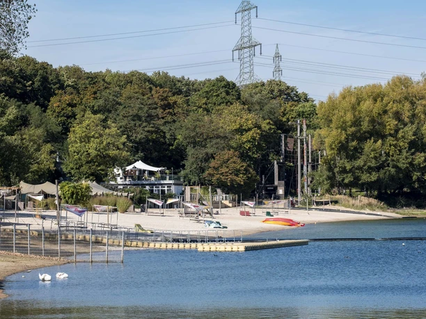 Blackfoot Beach Beach on Fühlinger See in front of a green forest, with an adjacent café and colorful boats on the water.