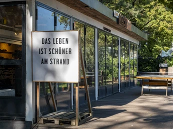 Blackfoot Beach Holzterrasse mit Strandbar und Tischtennisplatte, umgeben von Bäumen und Sonnenschein.Wooden terrace with beach bar and table tennis table, surrounded by trees and sunshine.