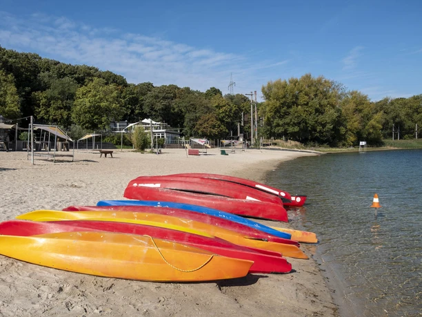 Blackfoot Beach Colorful kayaks on the sandy beach of a lake, surrounded by green trees under a blue sky.