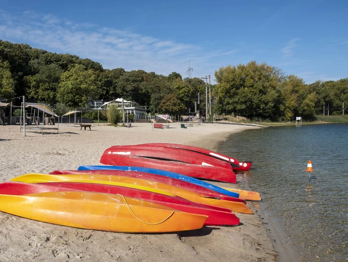 Blackfoot Beach Bunte Kajaks am Sandstrand eines Sees, umgeben von grünen Bäumen unter blauem Himmel.Colorful kayaks on the sandy beach of a lake, surrounded by green trees under a blue sky.
