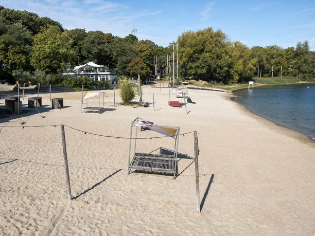 Blackfoot Beach A sandy beach with sunbathing areas, surrounded by a forest and clear water, under a blue sky.