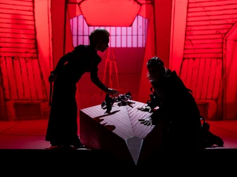 Zwei Darsteller auf einer dunklen Bühne, rot beleuchtet, knien vor einem Sarg.