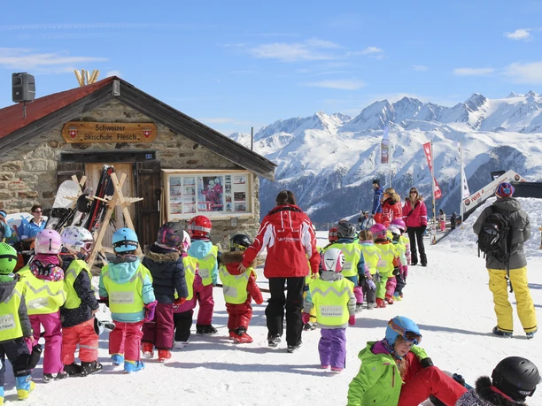 Cours de ski pour enfants à Fiescheralp Cours d'initiation au ski Fiescheralp avec monitrice de ski et enfants devant l'École Suisse de Ski de Fiesch par temps ensoleillé