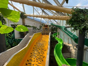 AQUALAND - wellness and aquapark Beleuchtete Indoor-Wasserrutschen, umgeben von tropischen Pflanzen und Sonnenschein durch Glasdach.Illuminated indoor water slides, surrounded by tropical plants and sunshine through the glass roof.