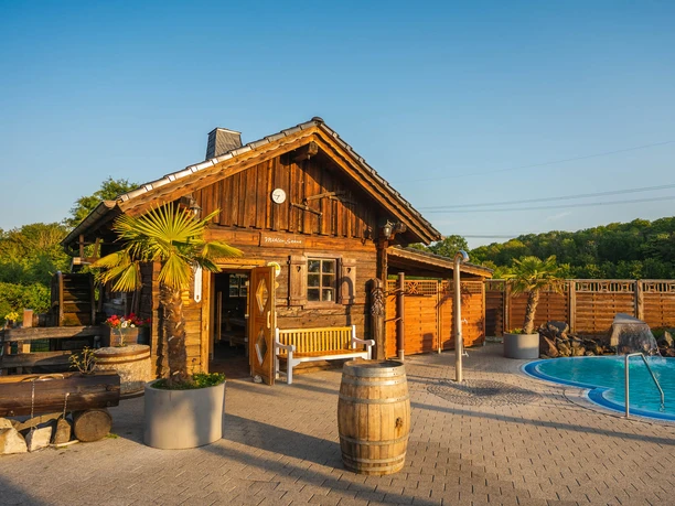 AQUALAND - Wellness und Erlebnisbad Ein rustikales Holzhaus steht neben einem runden Außenpool, umgeben von grüner Landschaft.