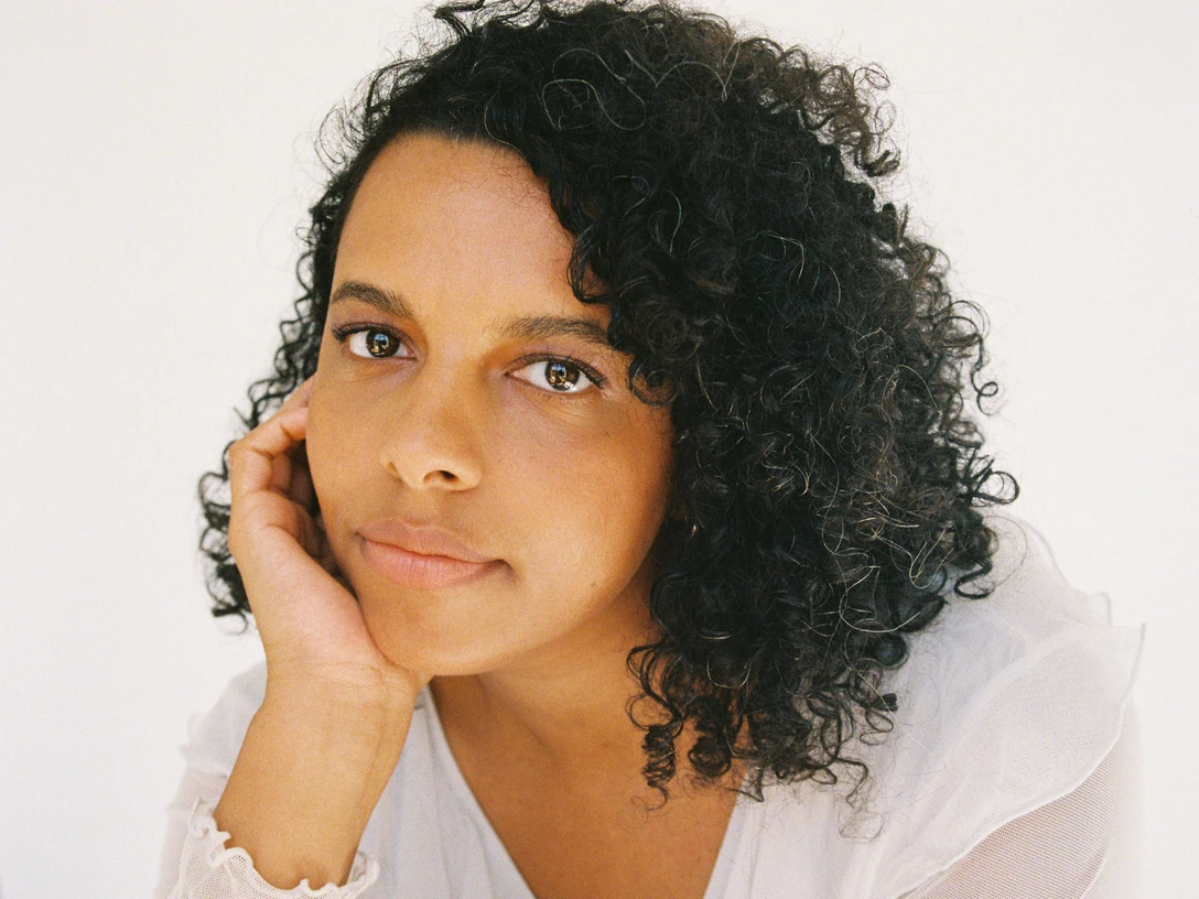 Press photo Alice-Hasters Das Foto zeigt die Künstlerin Alice Hasters im Portrait. Sie hat dunkle lockige Haare und trägt ein weißes TShirt.The photo shows a portrait of the artist Alice Hasters. She has dark curly hair and is wearing a white T-shirt.De foto toont een portret van de kunstenares Alice Hasters. Ze heeft donker krullend haar en draagt een wit T-shirt.