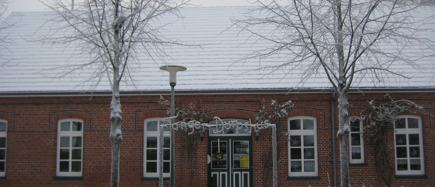 Brick building with white windows in winter, snow-covered trees and a hedge in front of it.