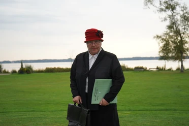 Fräulein Lisbeth Müller Ältere Frau mit rotem Hut und schwarzer Kleidung steht auf einer Wiese vor einem See im Hintergrund.