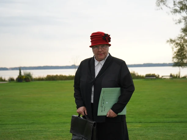 Fräulein Lisbeth Müller Ältere Frau mit rotem Hut und schwarzer Kleidung steht auf einer Wiese vor einem See im Hintergrund.