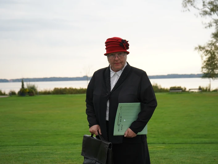 Fräulein Lisbeth Müller Ältere Frau mit rotem Hut und schwarzer Kleidung steht auf einer Wiese vor einem See im Hintergrund.
