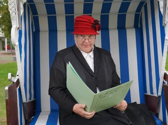 Fräulein Müller im Strandkorb Ältere Frau mit rotem Hut sitzt im blau-weißen Strandkorb und hält eine geöffnete Mappe.