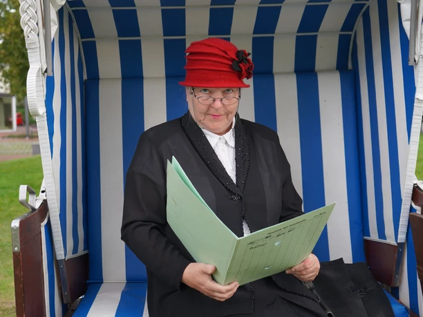 Fräulein Müller im Strandkorb Ältere Frau mit rotem Hut sitzt im blau-weißen Strandkorb und hält eine geöffnete Mappe.