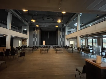 IMG_9236.jpg Großer Veranstaltungssaal mit Bühne, Holzboden, Tischen und Stühlen in moderner Industrieoptik.