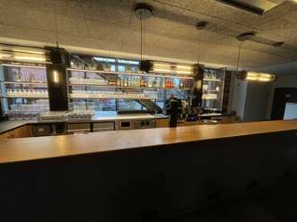 IMG_9237.jpg Moderne Bar mit Holztheke, beleuchteten Glasregalen, Getränkeflaschen und Kaffeemaschine.