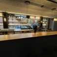 IMG_9237.jpg Moderne Bar mit Holztheke, beleuchteten Glasregalen, Getränkeflaschen und Kaffeemaschine.